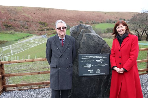 Prif Weinidog Cymru a'r Gweinidog yr Amgylchedd yn sefyll wrth ymyl plac llechen ar ddarn mawr o garreg o chwarel leol, gyda'r arglawdd llifogydd a chwm Dulais y tu ôl iddynt