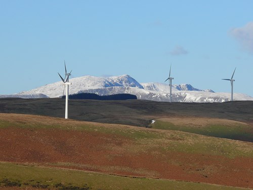 Gwaun ucheldir yn y blaendir gyda thyrbinau gwynt a mynyddoedd wedi’u gorchuddio ag eira yn y cefndir yn erbyn yr awyr las, glir